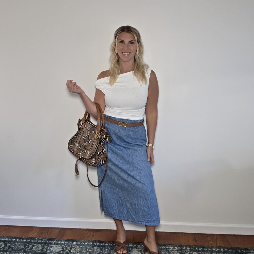 Woman over 40 in a white top and denim maxi skirt with a brown belt and printed shoulder bag