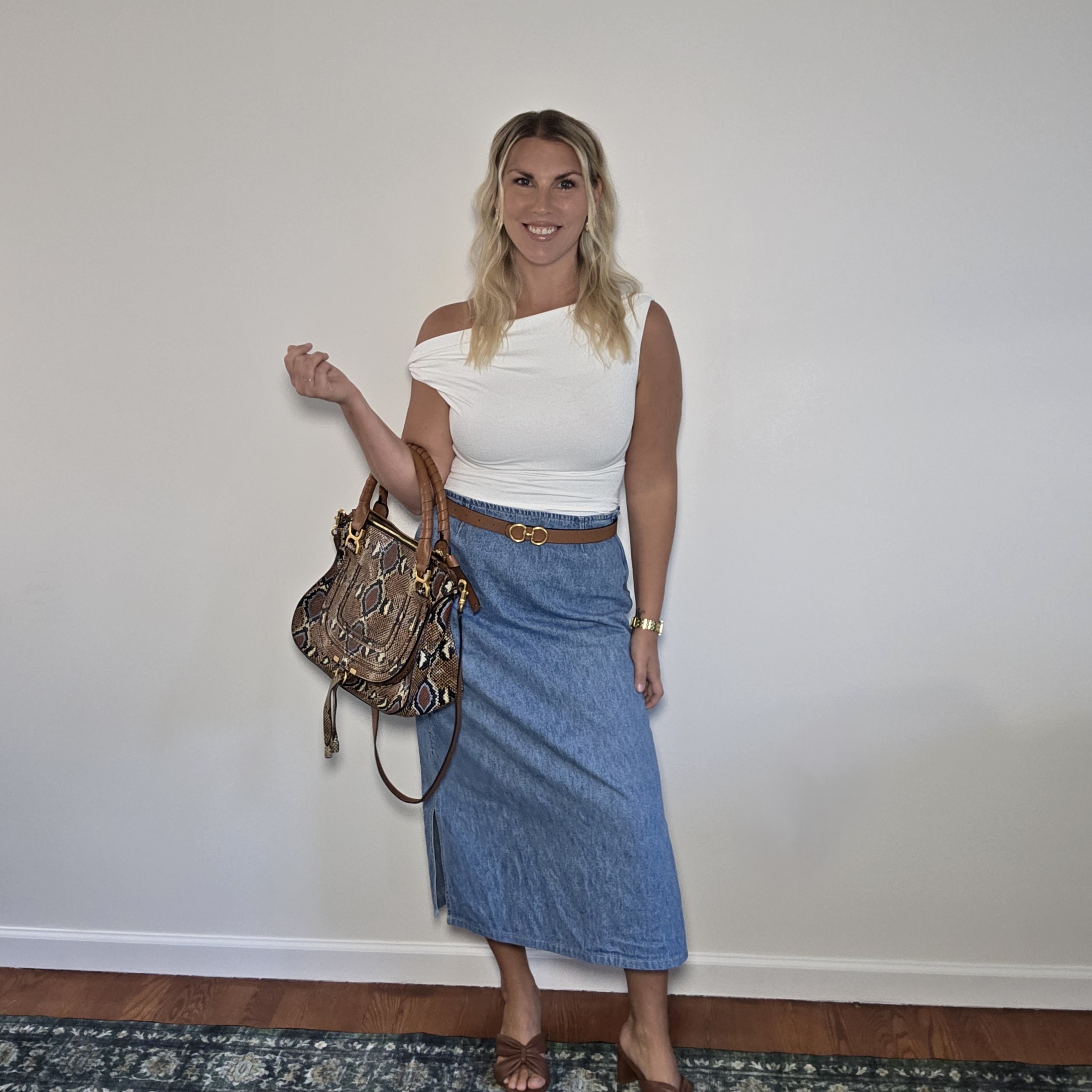 Woman over 40 in a white top and denim maxi skirt with a brown belt and printed shoulder bag