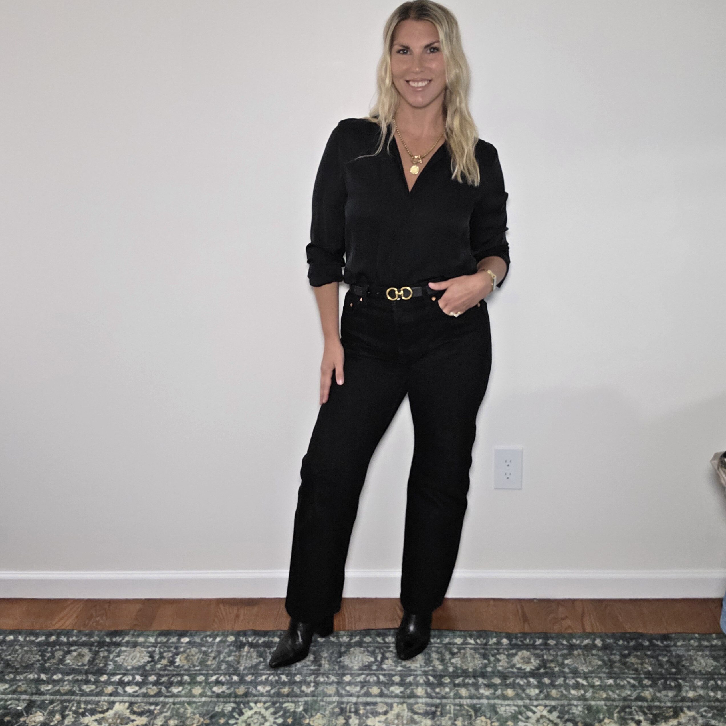 Tara wearing a black V-neck blouse tucked into black Levi's 501 straight-leg jeans with a gold-buckle belt and layered gold necklaces