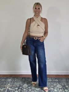 Woman over 40 in a tan halter wrap top and dark wide-leg jeans with a brown bag