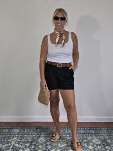 Woman over 40 in a white tank, black tailored shorts and brown belt with a straw bag