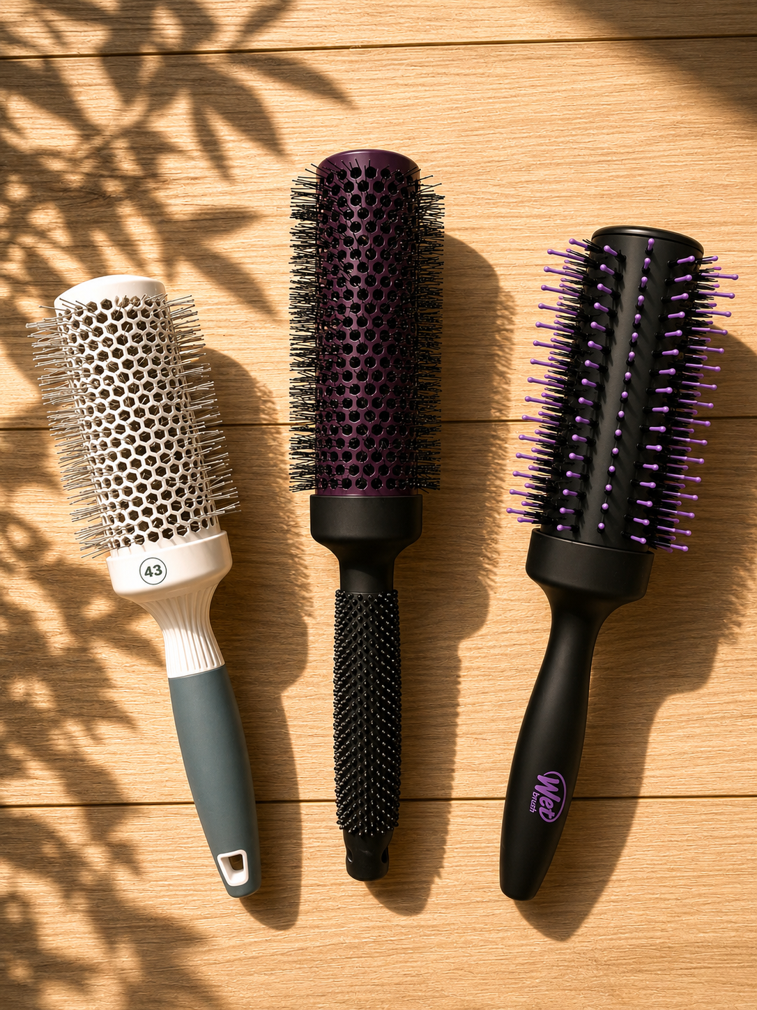 Three round barrel blowout brushes in cream, plum and black lined up on a wood surface in dappled sunlight
