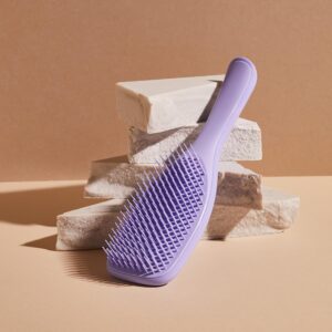 Lavender detangling hair brush resting against textured stones on a tan backdrop