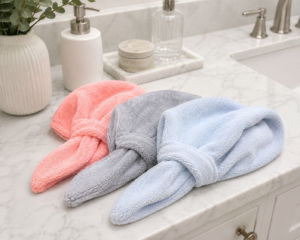 Three twisted microfiber hair towels in coral, gray, and light blue on a white marble bathroom counter