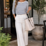 Woman over 40 in a navy-and-white Breton striped top, white wide-leg trousers and tan loafers, carrying a cream tote outside a cafe