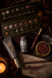 Moody flatlay of vamp-romantic fall makeup products — a berry eyeshadow palette, foundation and highlighter with dark roses